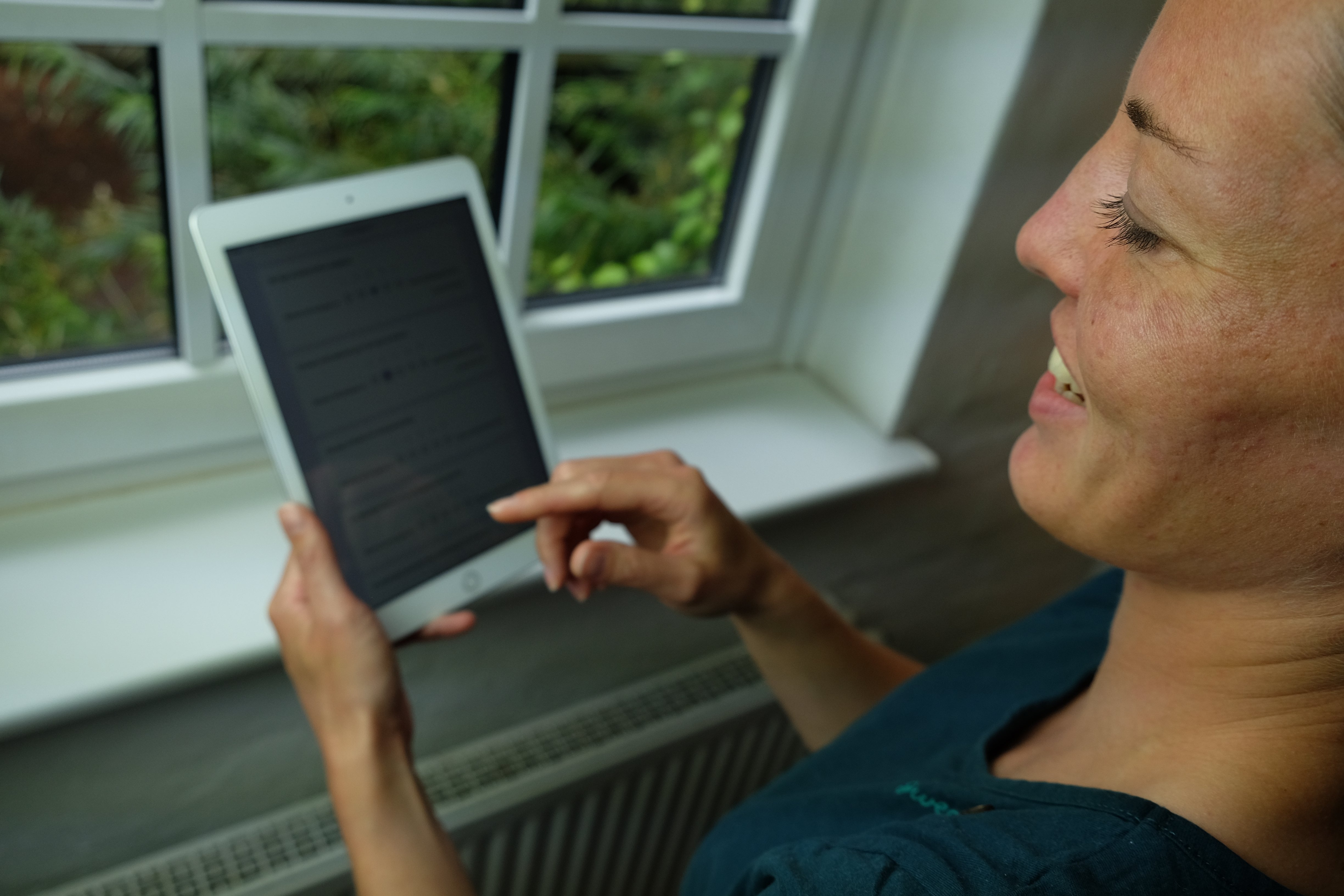 Person steht lächelnd vor einem Fenster und hat ein Tablet in der Hand auf das sie tippt.